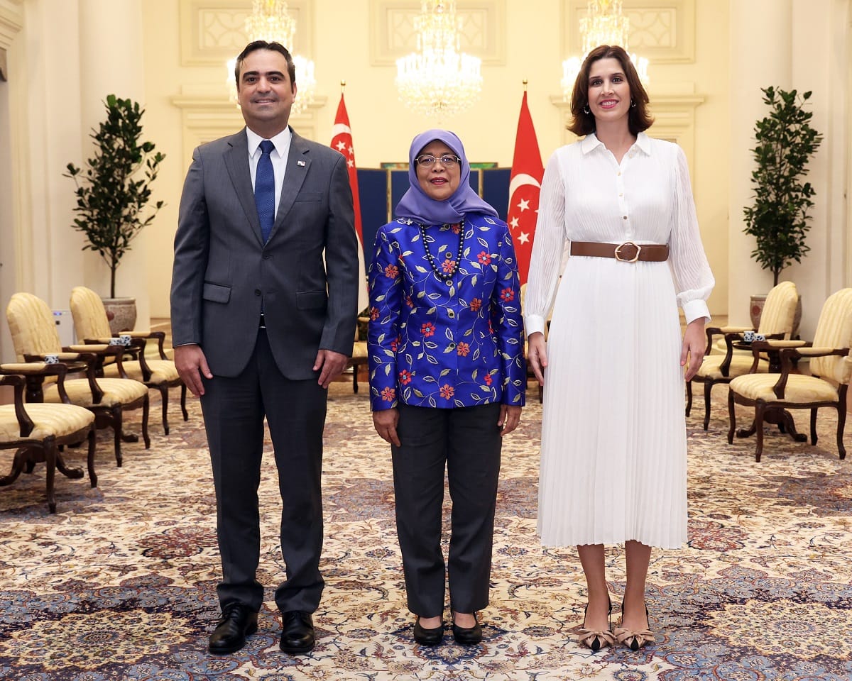 Three people stand in a room with Singapore flags and ornate furnishings.